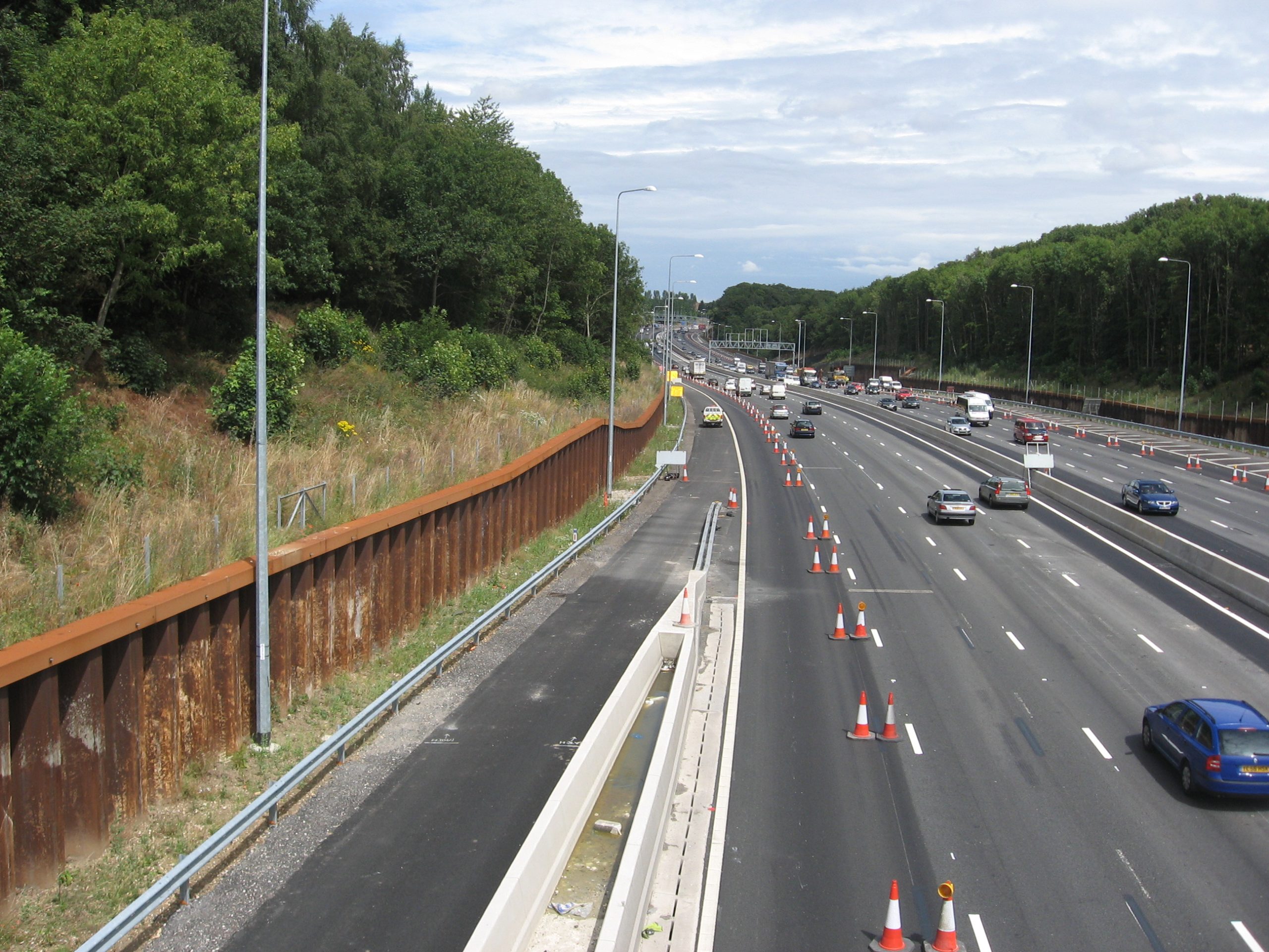 Steel sheet piling retaining wall installed along the edge of a dual carriageway motorway with traffic cones and vehicles in motion